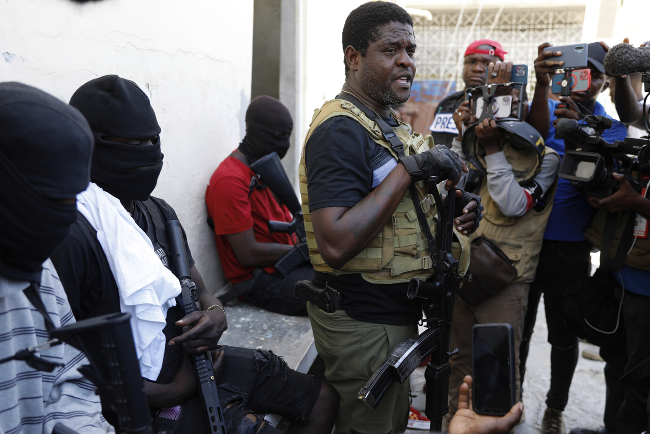 a man weaing a bullet proof vest speaks to a group of men who are recording him with their smartphones. three men with black ski masks sit on either side of the standing man.