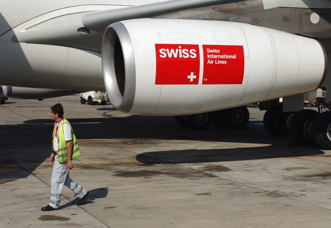 A man in a high visibility jacket is walking on the runway tarmac alongside the engine of a white SWISS plane. The SWISS logo is visible on the engine: SWISS in white lettering on a red backdrop with the Swiss flag 'plus' in the corner, and the full acronym: Swiss International Air Lines