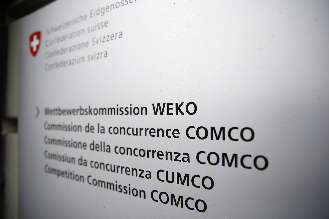 A white sign with the Swiss Confederation red shield with a white plus in the top left-hand corner. The sign says ‘Competition Commission COMCO’ in Switzerland’s four different languages as well as English
