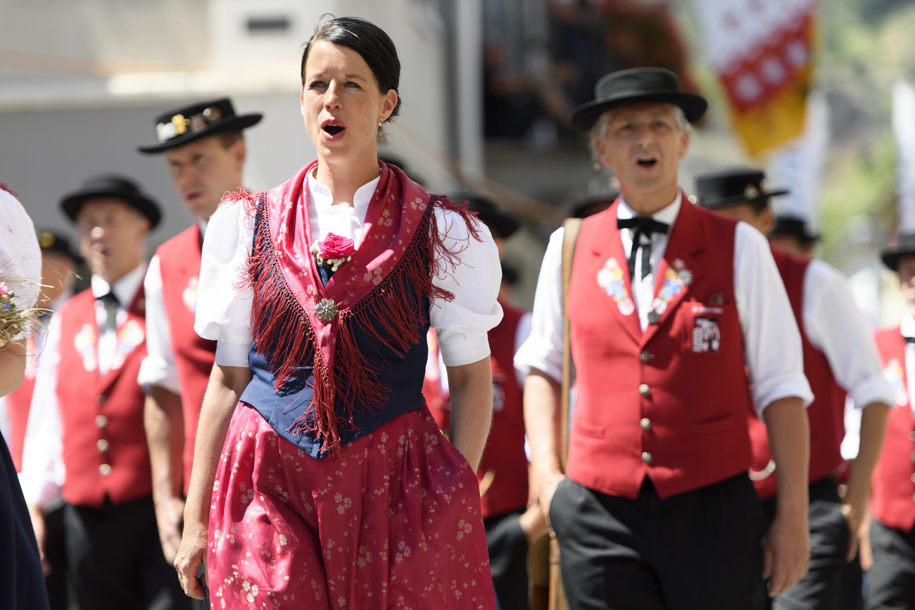 Yodellers walk through the streets of Brig for the Yodelling Festival in traditional costume. A dark-haired woman yodelling is at the forefront of the photo in a white, short-sleeved blouse, navy waistcoat and red scarf and skirt. Behind her are men yodelling in black hats, white shirt sleeves, red waistcoats and black trousers.