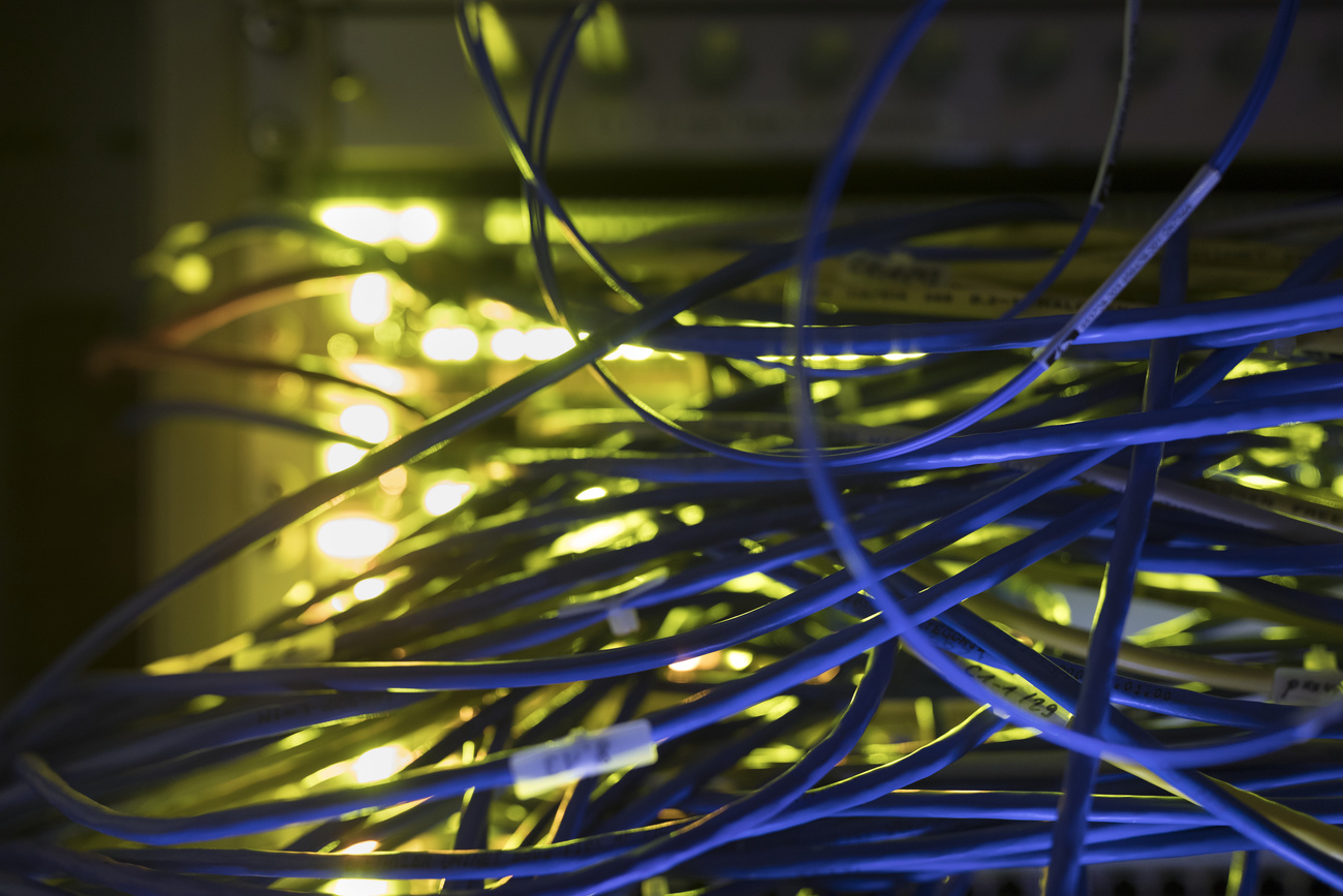 Patch panels, LED, fibre optic cables and switches in a server room. A tangle of blue cables is visible at the foreground of the photo with yellow lights in the background.
