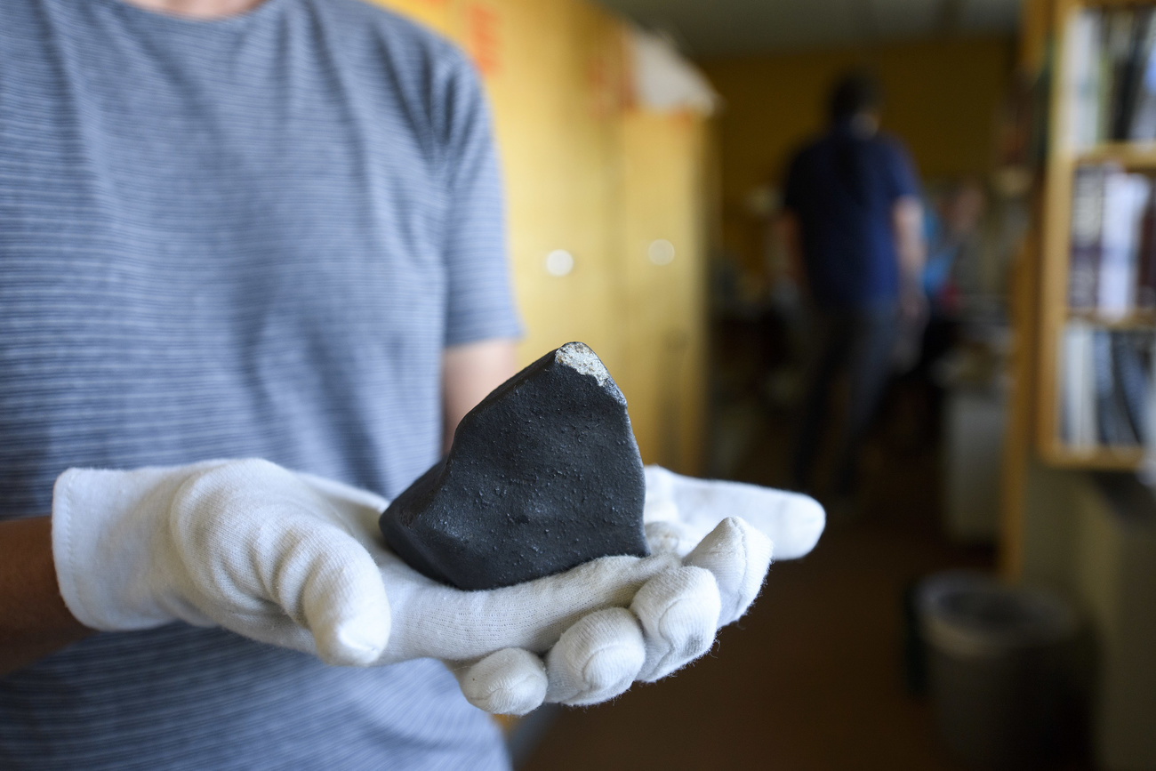 A person in a light blue and white striped T-shirt and white cotton gloves holds out a smooth black meteorite to the camera