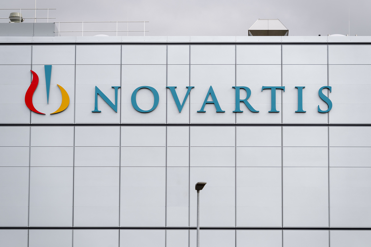 A plain white factory building with the Novartis logo on the side: ‘Novartis’ in turquoise text and on the left three lines resembling a mortar and pestle — two smooth strokes in dark red and orange curve around a straight vertical turquoise line.