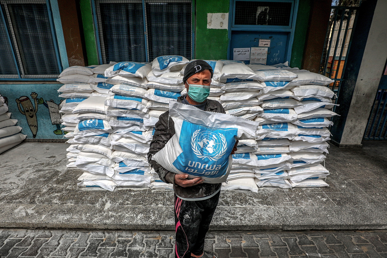 A man in a dark cap, jacket and black trousers with red stripes down the side, holds a white food aid sack with the blue UNRWA logo on the front. He is wearing a blue protective face mask and behind him are piles of the same sacks.