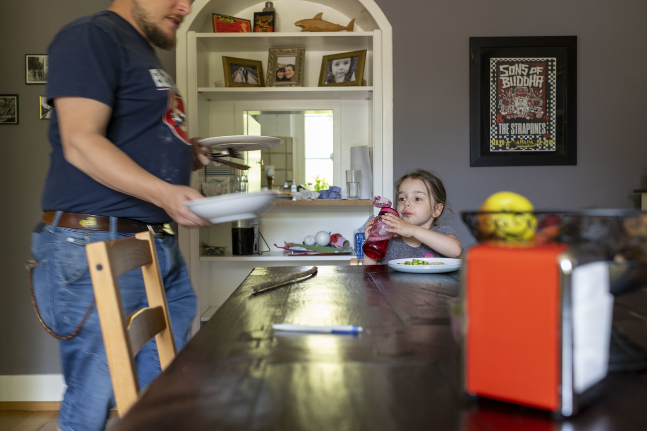 man wearing a blue t-shirt and jeans gives young girl a plate of food on a white plate while she drinks from a red sippy cup