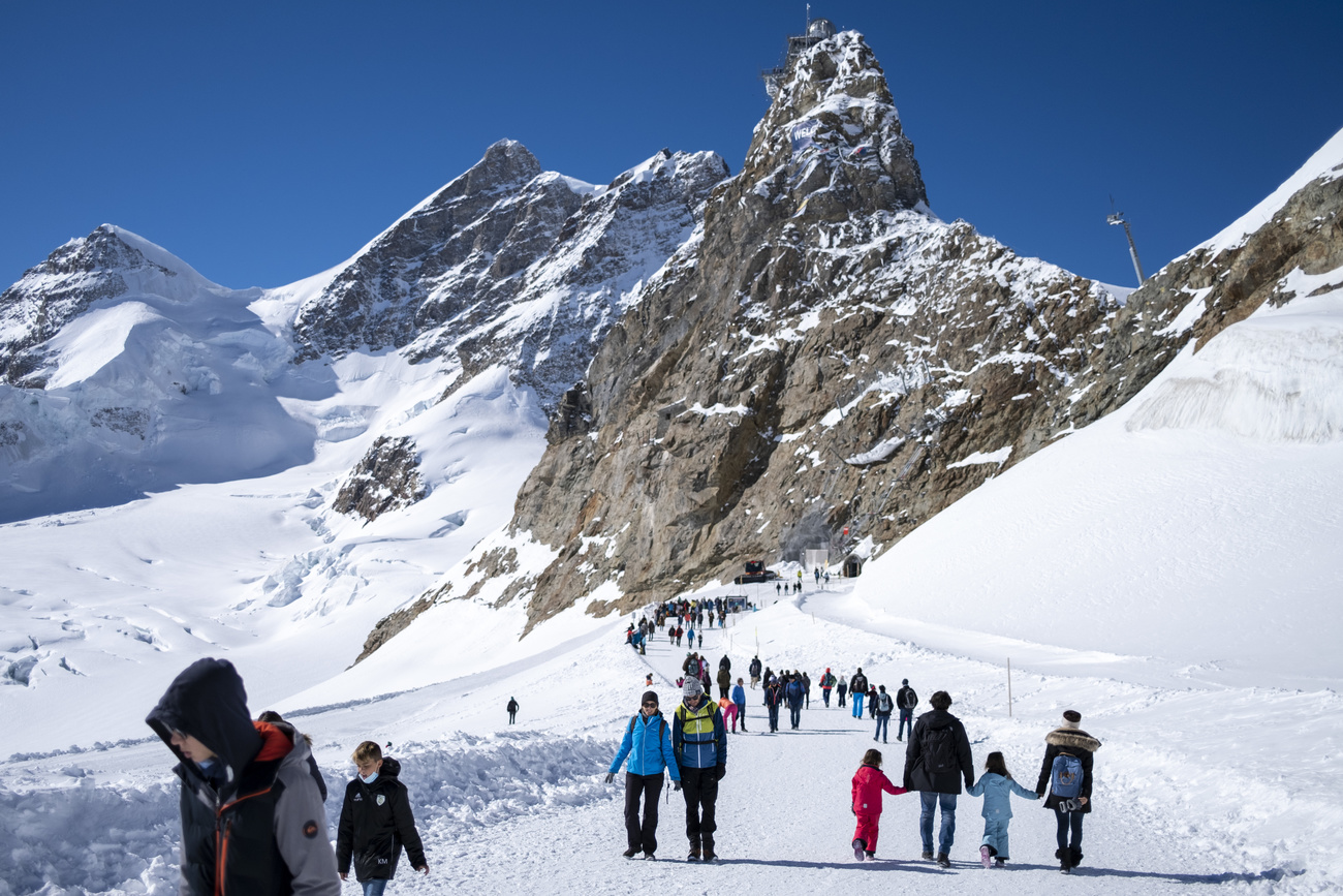 Tourists walk along a snowy path, behind them is the Jungfraujoch mountain peak and station