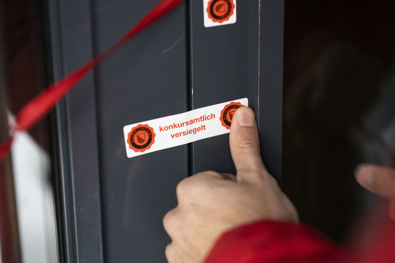 A white bankruptcy office sticker is being placed over the opening of a door, which says ‘konkursamtlich versiegelt’ in red letters, which means ‘sealed by the bankruptcy authorities’