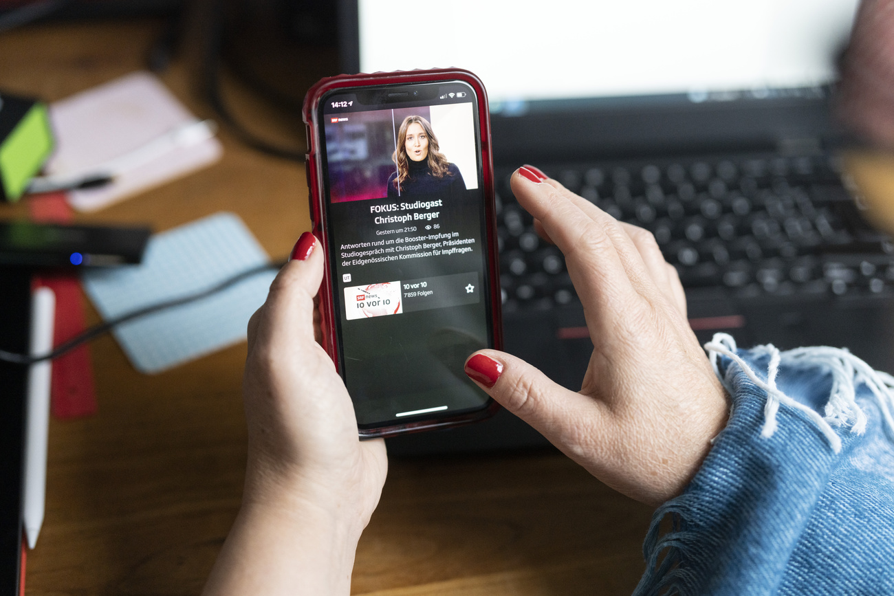 A picture of a young woman’s hands, with red painted nails. She is using the SRF app on her phone, which has a red case. Blurred in the background on her desk are a pen, post-its and her laptop.