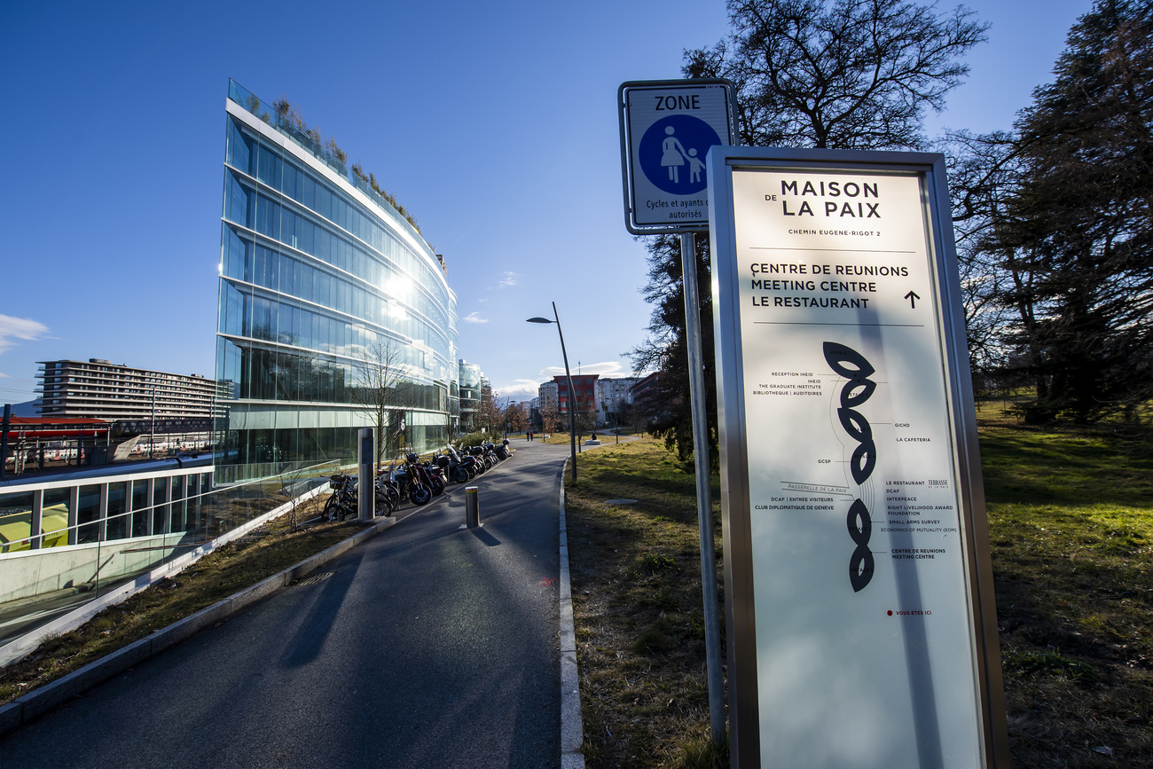 A white sign says ‘Maison de la Paix’ to the right of a pedestrian walkway. Behind it to the left is a glass building, to the right is a grassy area.
