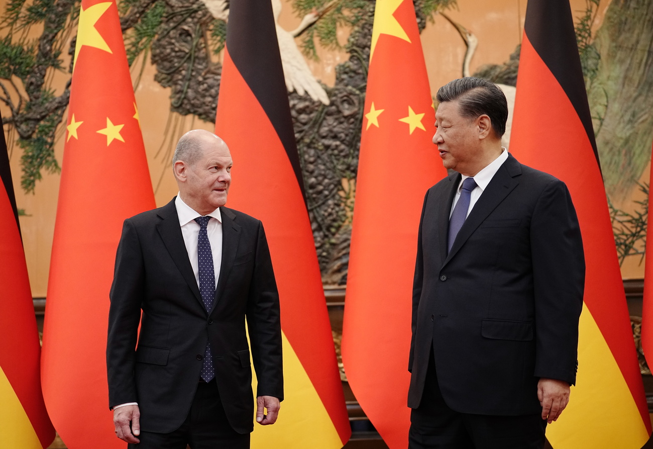 President of China Xi Jinping, right, and German Chancellor Olaf Scholz, left, stand facing each other and are speaking together, although their bodies are turned outwards towards the camera. Both are wearing dark ties, white shirts and black suits. Behind them are alternating Chinese and German flags – the German flag with a black stripe on top, then red, then yellow, while the Chinese flag is red with a big yellow star in the top left-hand corner and a circle of yellow stars around it.