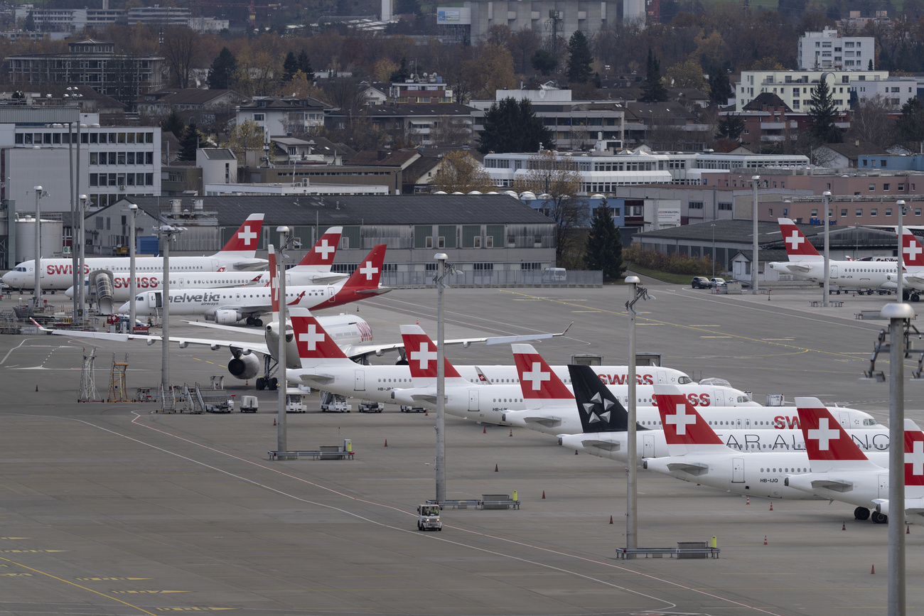 Eight white SWISS planes parked on the tarmac at Zurich airport. Their red tailfins have the white Swiss flag ‘plus’.