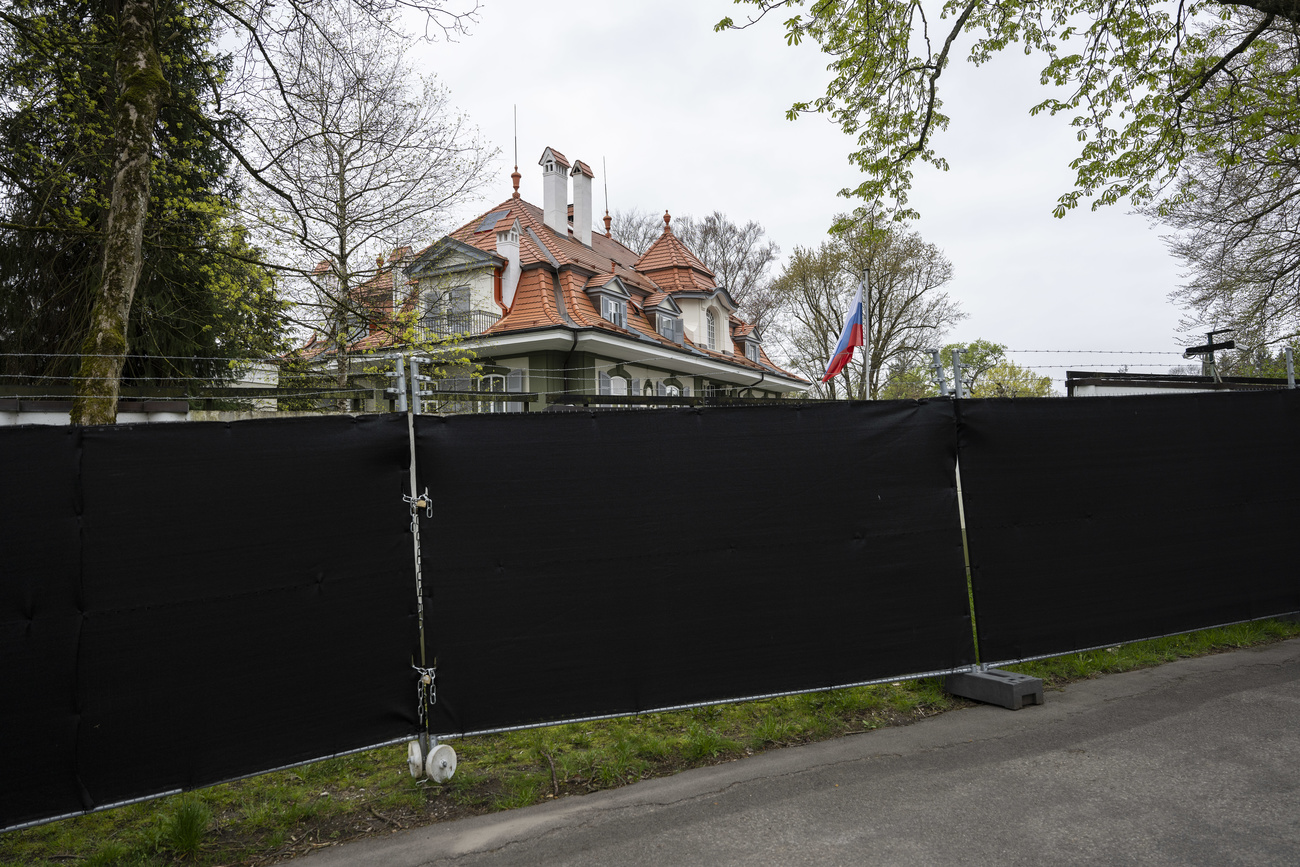 Russia’s embassy building in Switzerland: a white building with a terracotta roof and a Russian flag flying outside. The building is behind a large black barrier that blocks most of the view of the building.