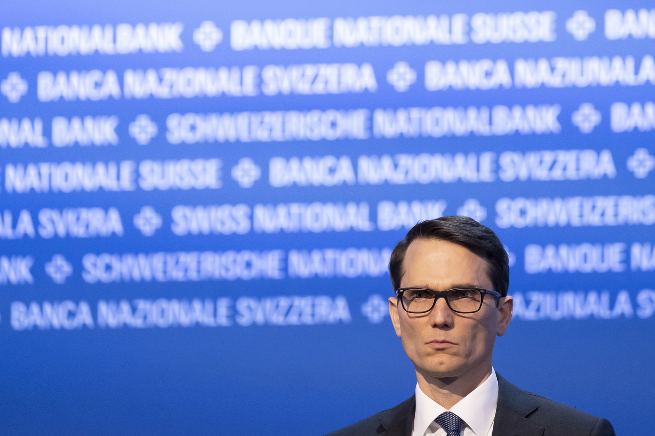 Man in black suit wearing thick rimmed black glasses stands in front of blue Swiss National Bank wall