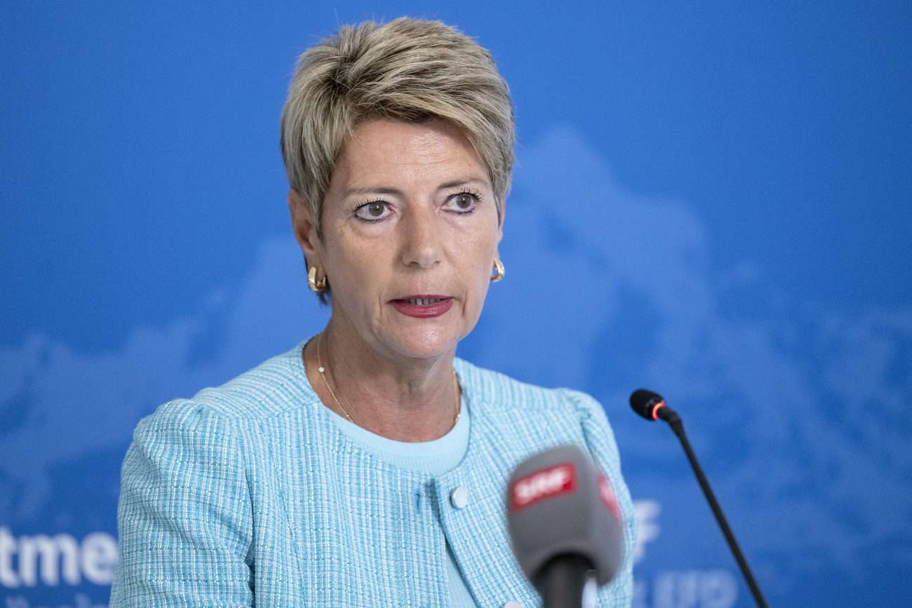 Swiss Finance Minister Karin Keller-Sutter speaks into two microphones. She has short blonde hair and is wearing a turquoise jacket and gold hoop earrings. Behind her is a light blue background.