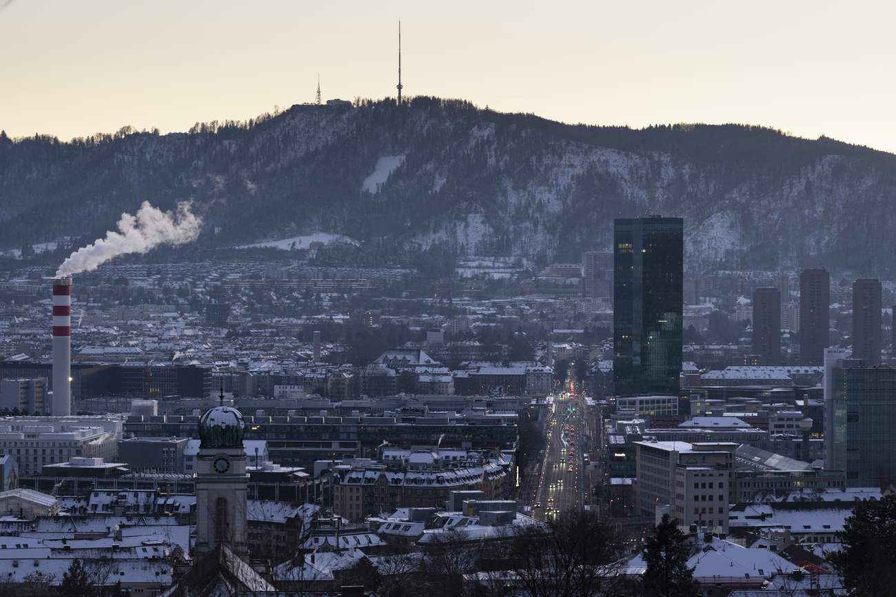A view across Hoengg in Zurich including Hardbruecke, the Prime Tower and the Uetliberg, at nightfall. Roofs and the mountain are covered in snow.