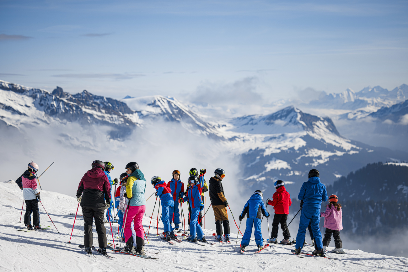 Groups of skiers in brightly coloured outfits gather at the top of a snow-covered piste with white mountains visible in the distance