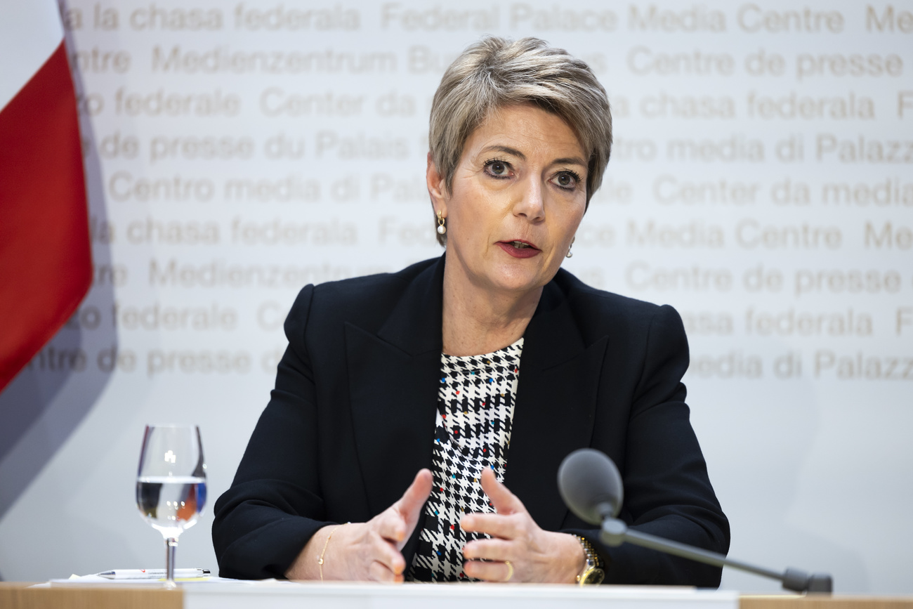 Karin Keller-Sutter sits at a wooden desk with a glass of water to her right. The red Swiss flag with the white plus can be seen behind her as well as the words ‘media centre’ in various languages on the white wall. She has short hair and is wearing a black jacket and a monochrome houndstooth patterned outfit. She has her hands out open in front of her with the sides of her palms resting on the table, and is speaking into a microphone.