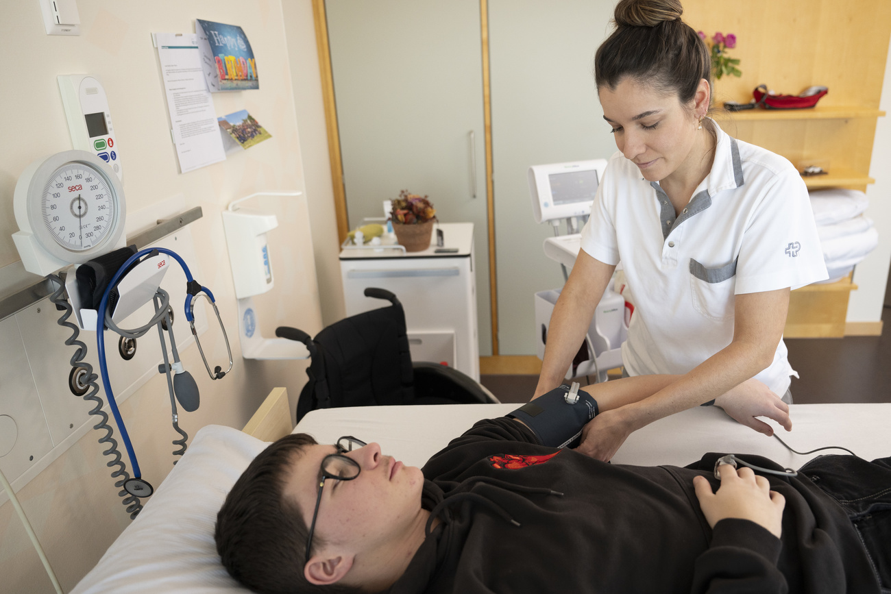 A nurse with her dark hair tied back in a bun and wearing a white shirt with grey trim on the collar and front pocket takes the blood pressure of a young man lying on a white medical bed. He is wearing a black jumper and glasses. The walls are light beige and as well as a white cabinet, medical devices such as a stethoscope, a white hand gel dispenser and a black manual wheelchair are visible in the room.