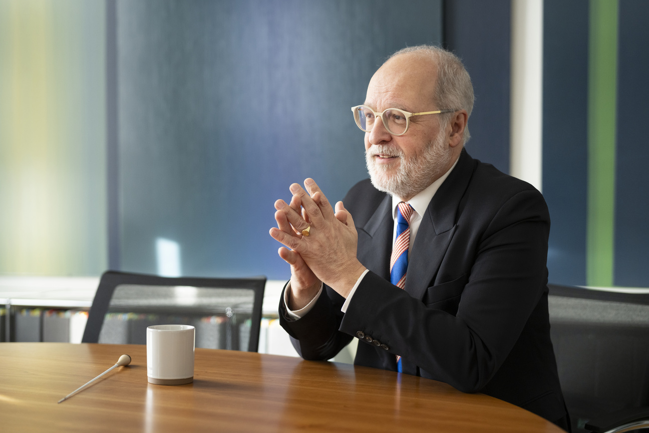 Federal Department of Foreign Affairs (FDFA) Secretary of State Alexandre Fasel sits with his elbows resting on a wooden table, a white mug is in front of him and his hands are together. He has a beard and wears glasses and a black suit, white shirt and a tie with alternating blue and red and white striped patches.
