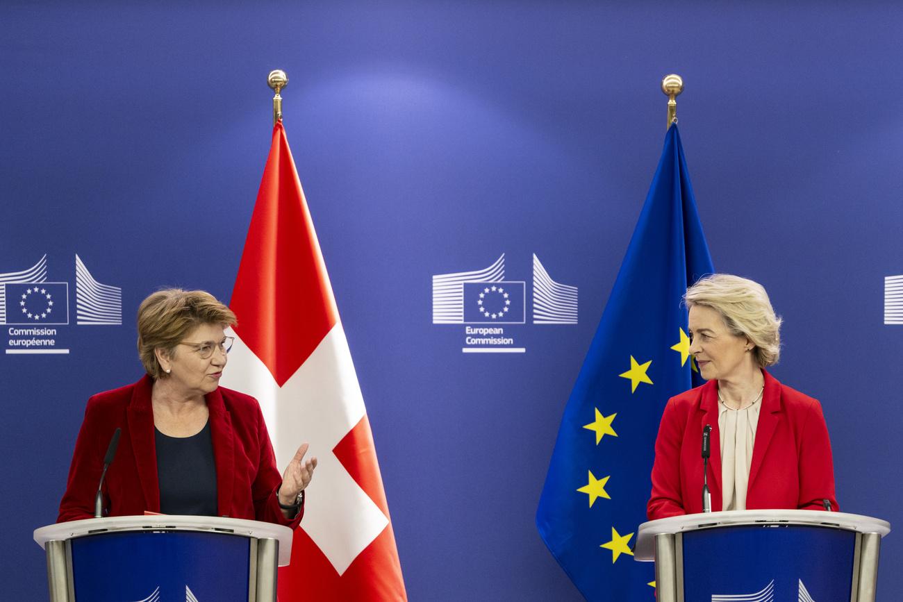 Swiss president Viola Amherd on the left, speaks to Ursula von der Leyen, president of the European Commission, on the right. Both stand in front of a blue European Commission backdrop behind their own podiums. Amherd wears a dark top and a red jacket and stands in front of the Swiss flag (a red square flag with a white plus). Von der Leyen wears a light top and a red jacket and stands in front of the EU flag, which is navy with a circle of 12 gold stars.