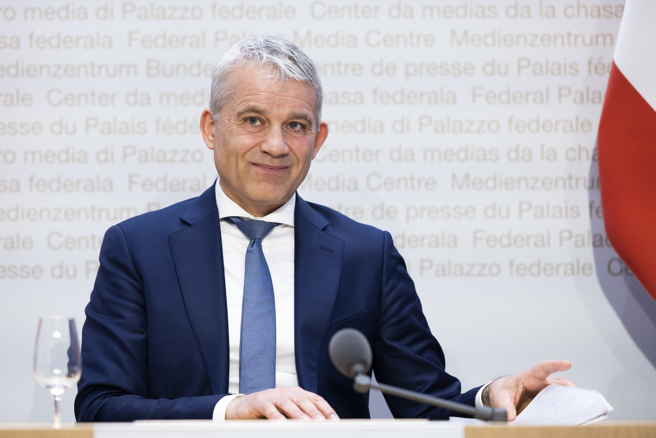 Justice Minister Beat Jans sits at a desk in the Federal Palace media centre, a glass to his right and a microphone in front of him. He is leafing through papers and wears a dark blue suit jacket, a blue tie, a white shirt and has short, white hair.