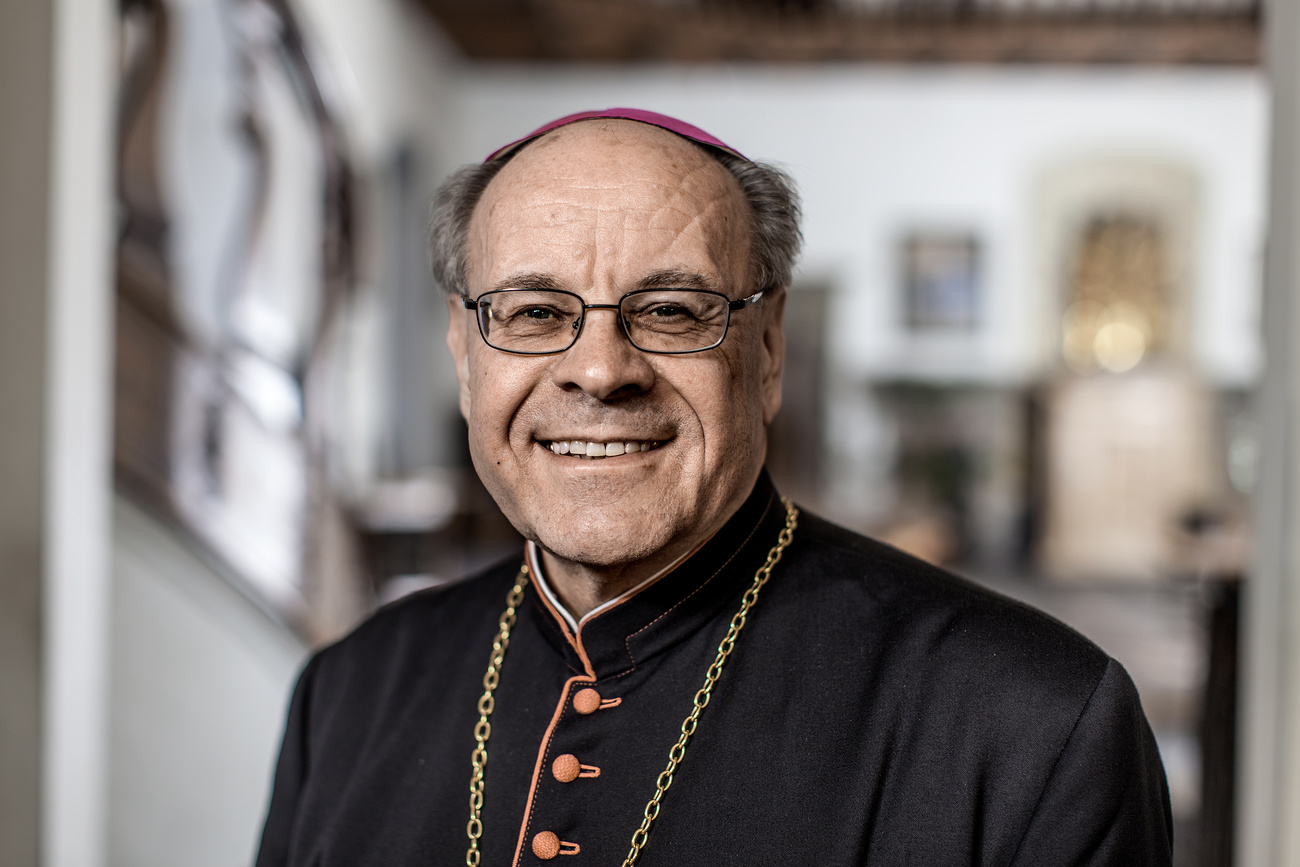 Vitus Huonder smiles into the camera. He has short grey hair under a dark red cap, glasses, and wears a black cassock with orange buttons and trim, and a golden chain around his neck.