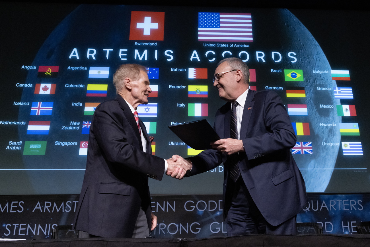 Swiss economics minister Guy Parmelin and NASA Administrator Bill Nelson, both wearing dark suits and white shirts, shake hands. Parmelin is holding the signed document in his left hand. In the background is a huge screen saying ‘Artemis Accords’, with the flags and names of all the other country signatories, with the Swiss and US flags at the top