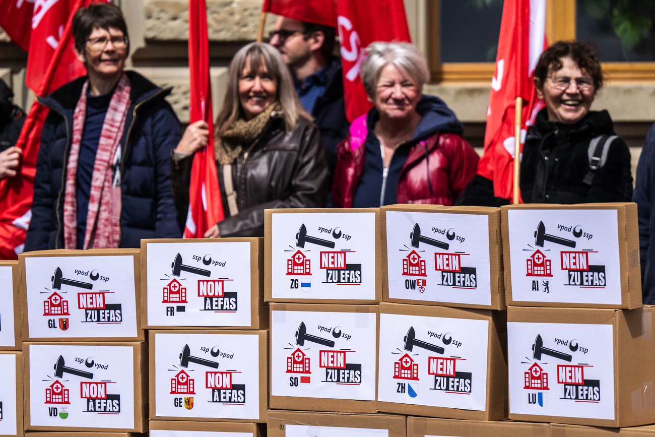 Four female protesters stand smiling with red flags outside parliament in Bern. In front of them are cardboard boxes with signs on that say ‘No to Efas’ with the public services union (VPOD) logo and a picture of a hammer hitting a hospital. At the bottom of each sign are the different cantonal flags.