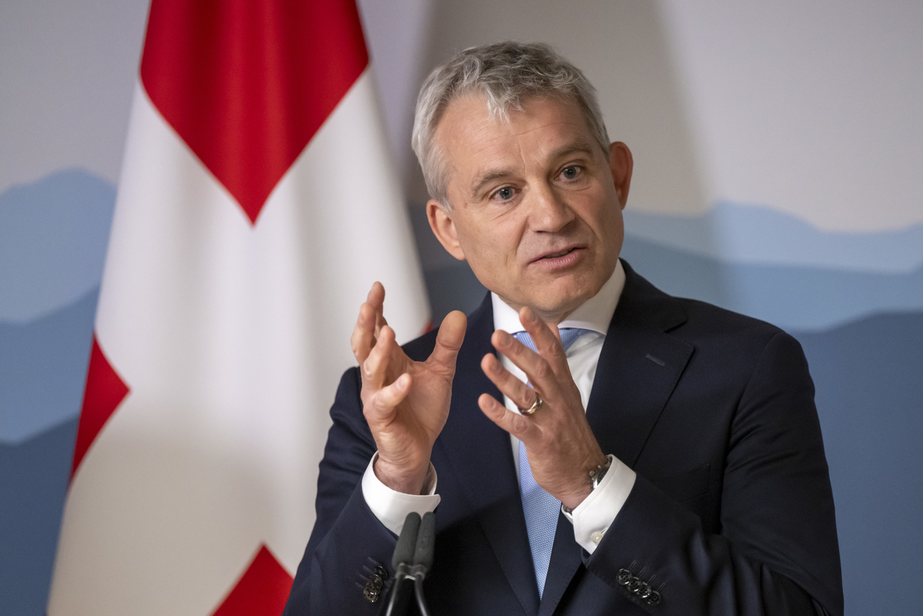 Justice Minister Beat Jans is speaking into a microphone with both hands raised. He is wearing a dark suit jacket, white shirt and a light blue tie. Behind him is a white wall depicted with blue mountains and a Swiss flag (red with a white plus).