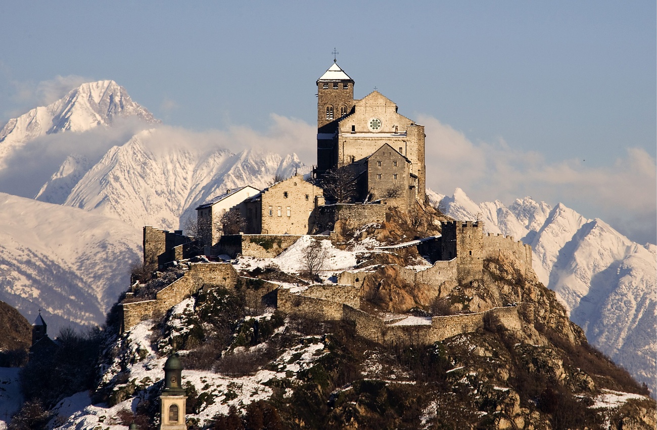 The 13th century Valère basilica, its walled enclosure and associated buildings perch on top of a snow-dusted hill. The snow-covered Bietschhorn mountain can be seen in the background.