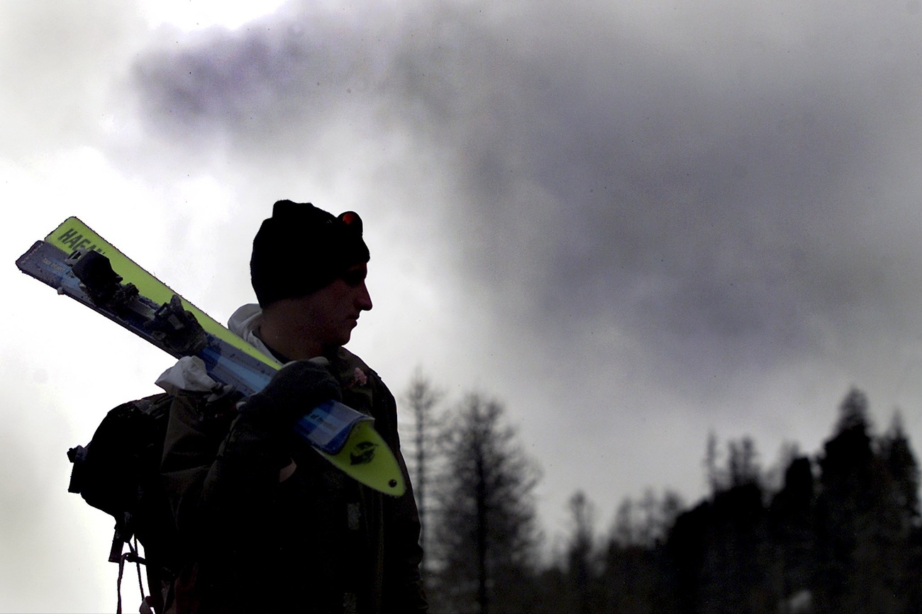 The dark outline of a competitor with his skis over his shoulder can be seen standing, with dark clouds behind him and the black outlines of trees