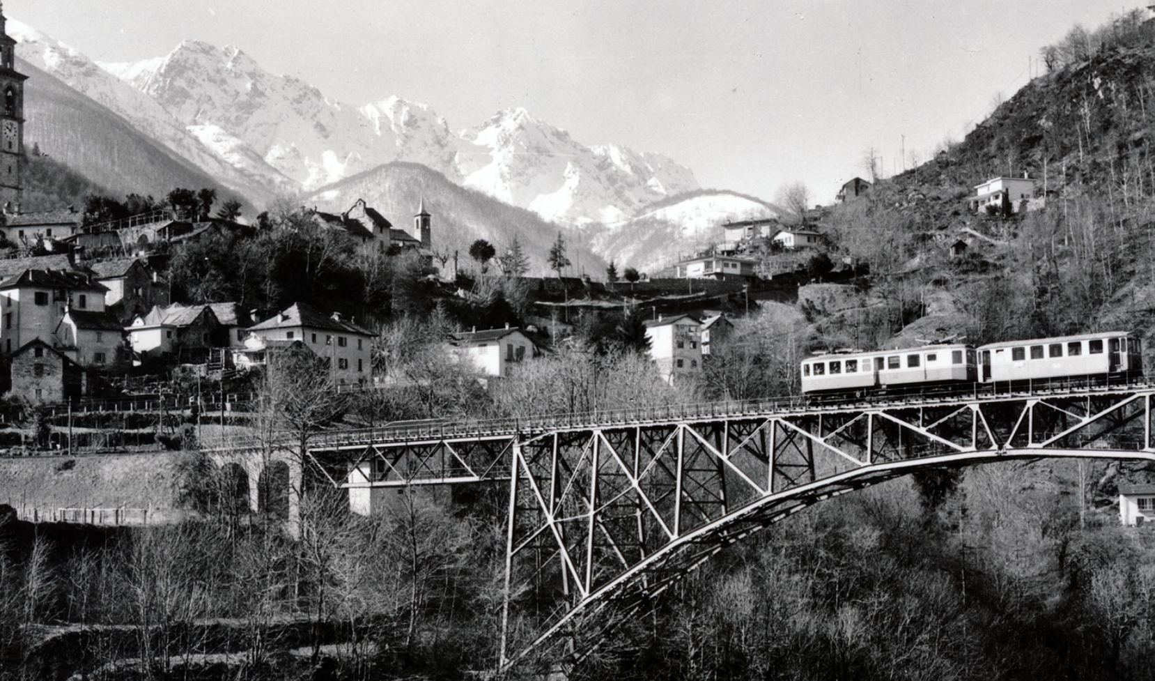 Motrice électrique passant un viaduc avec un village et des montagnes en arrière-fond.