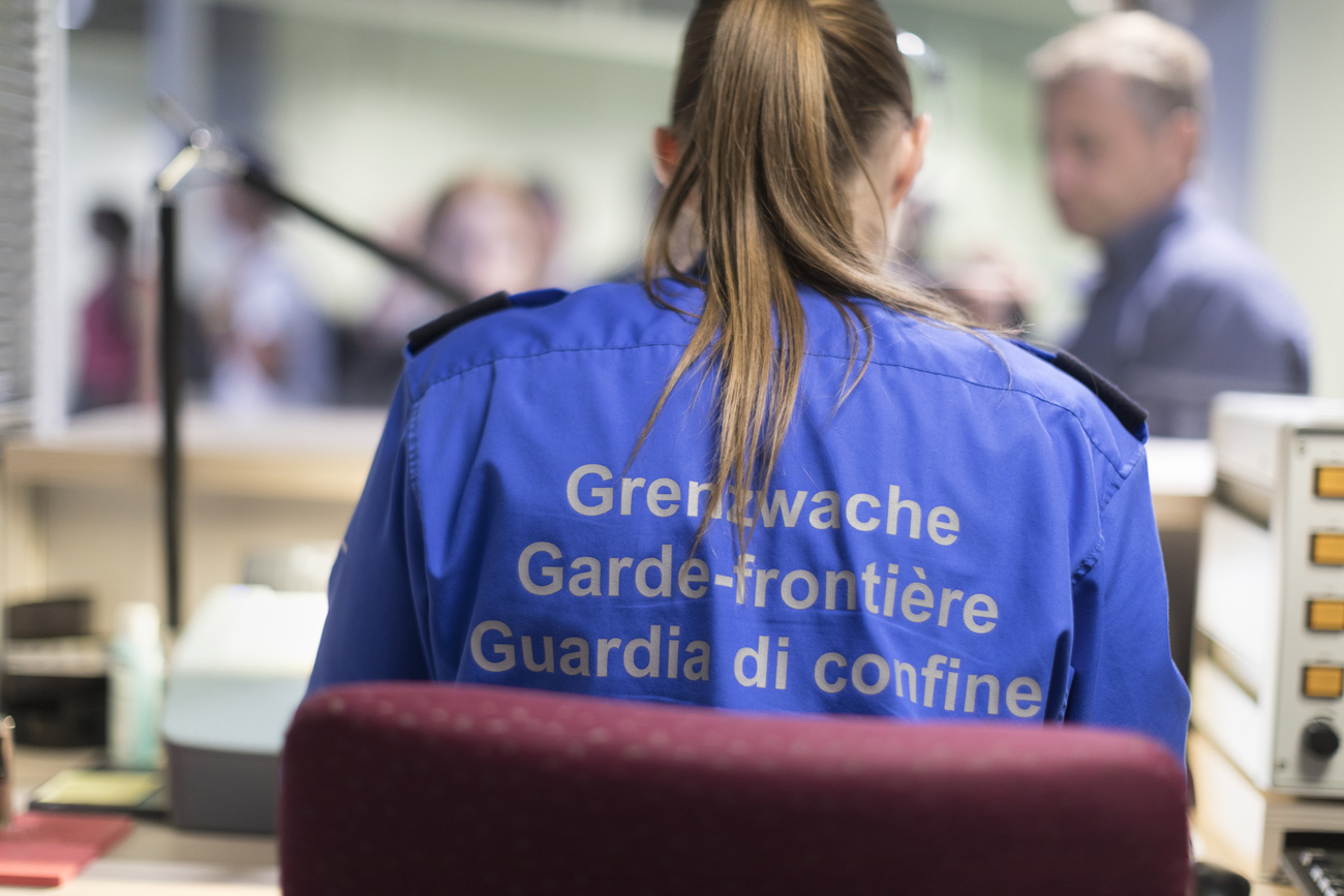 Pictured from behind, a female Swiss Border Guard employee is sitting on a burgundy chair. She is wearing a blue shirt that says ‘Border Guard’ on the back in at least three Swiss languages. Her long, dark blonde hair is tied into a neat ponytail.
