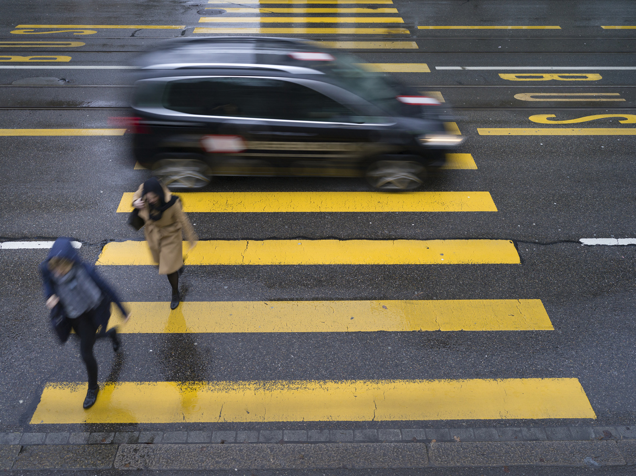 Two pedestrians, one in a blue coat, one in a beige mac, cross a yellow-striped pedestrian crossing on a road. Behind them, a black car drives over the crossing. They are blurred, the yellow crossing lines are in focus.