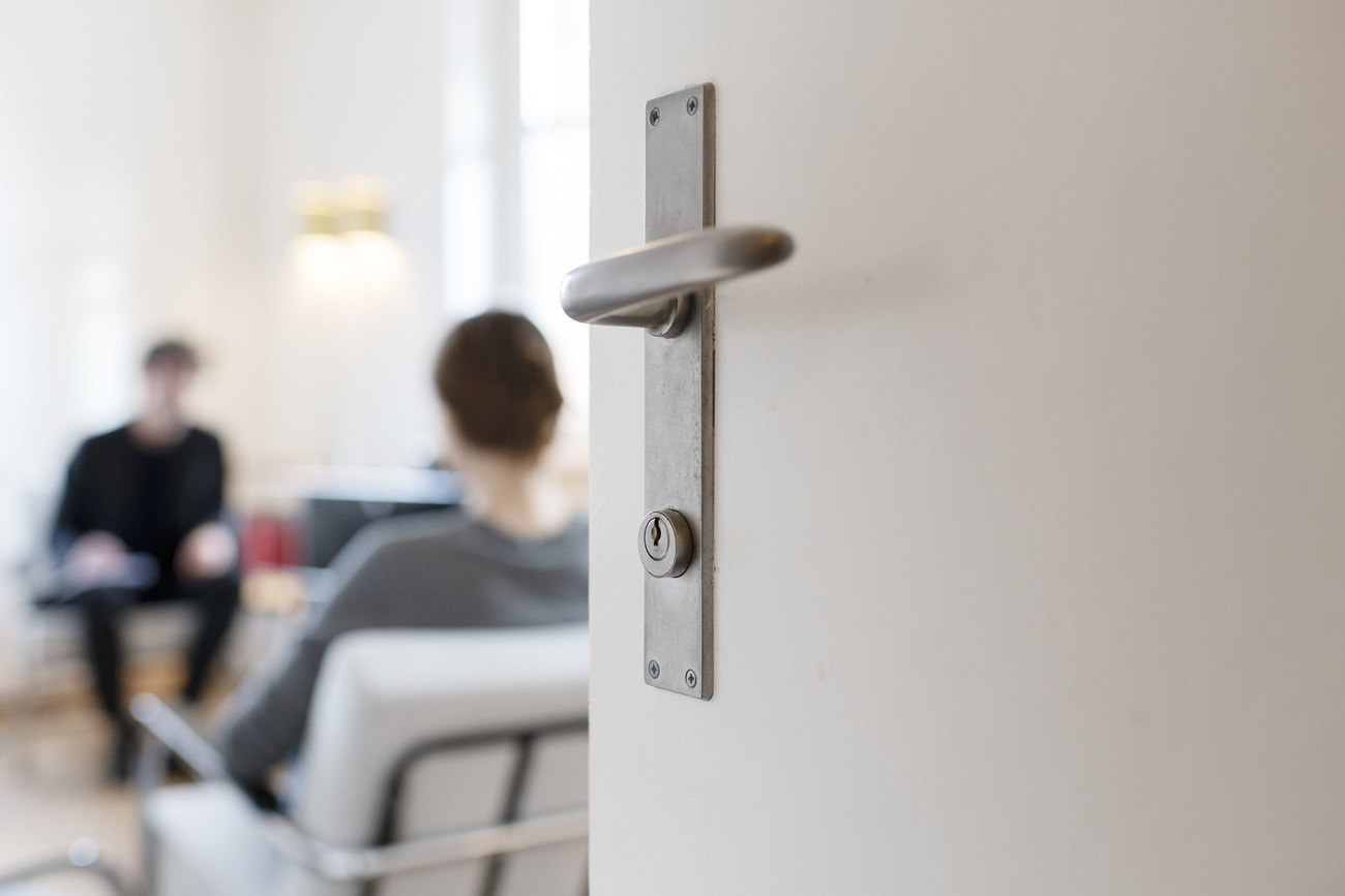 In the foreground is a white door with a grey metal door handle. In the background can be seen two people sitting opposite each other.