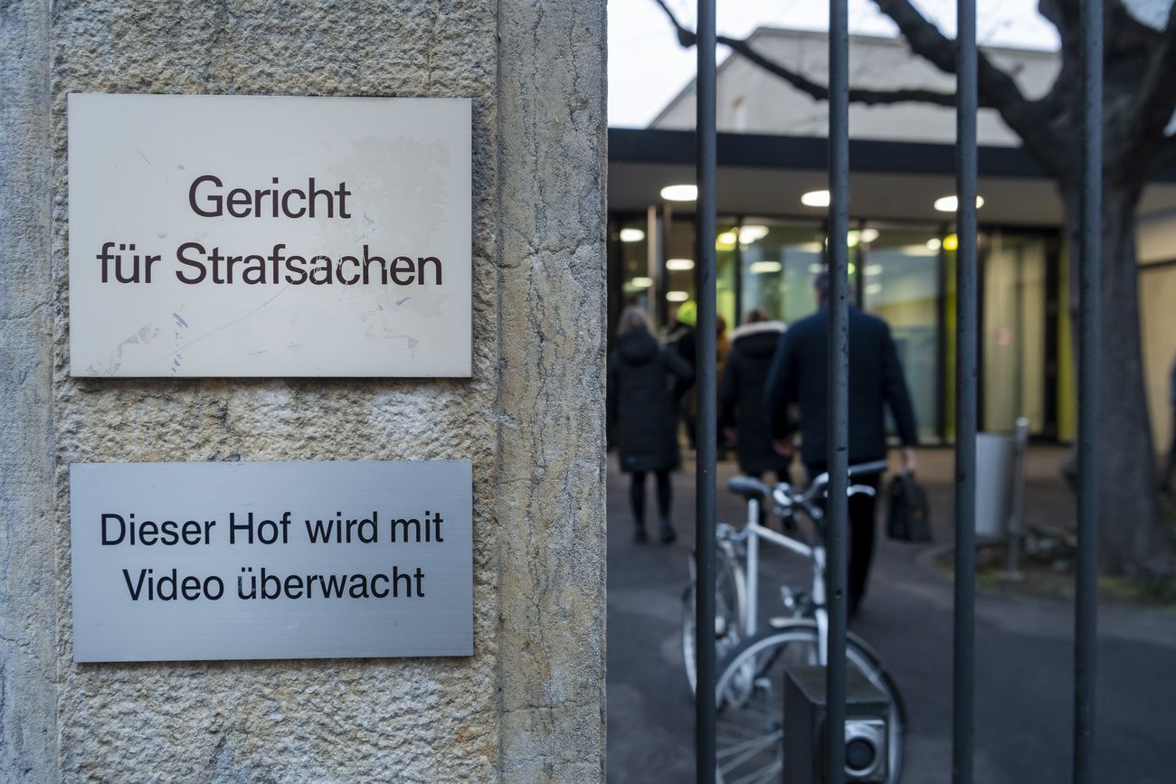 A view of the sign for the criminal court in Basel, with bars from a gate. Inside the gate, people can be seen walking towards the building.