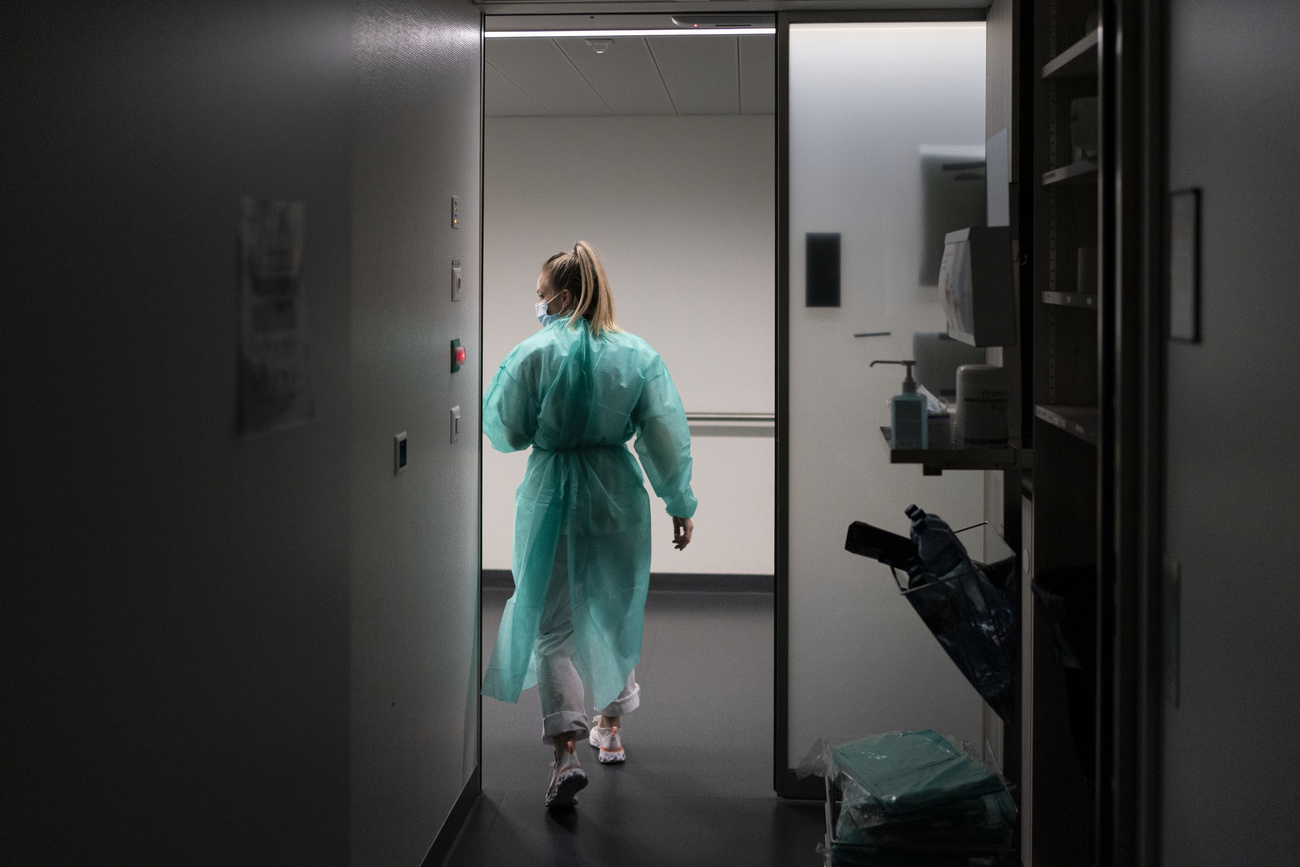 A female healthcare worker with a blonde ponytail and wearing a turquoise plastic coverall and face mask walks out of a dark room into a light hallway