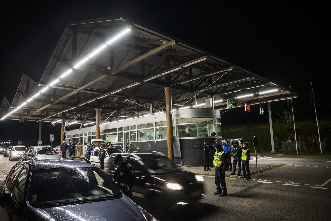 A number of cars are seen stopped at a border control station, with agents in uniform standing before them.