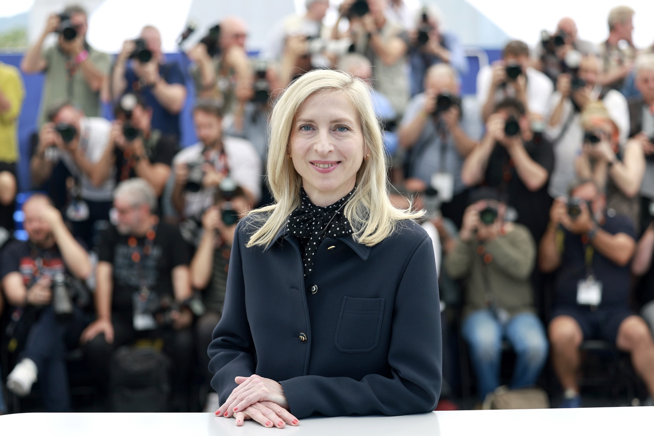 Austrian filmmaker Jessica Hausner pictured at the Cannes film festival. Her hands are on a white surface in front of her as she smiles into the camera. She has long blonde hair and wears a black coat with a white spotted black blouse undernearth with a bow tied at the neck. Blurred outlines of people are visible behind her.