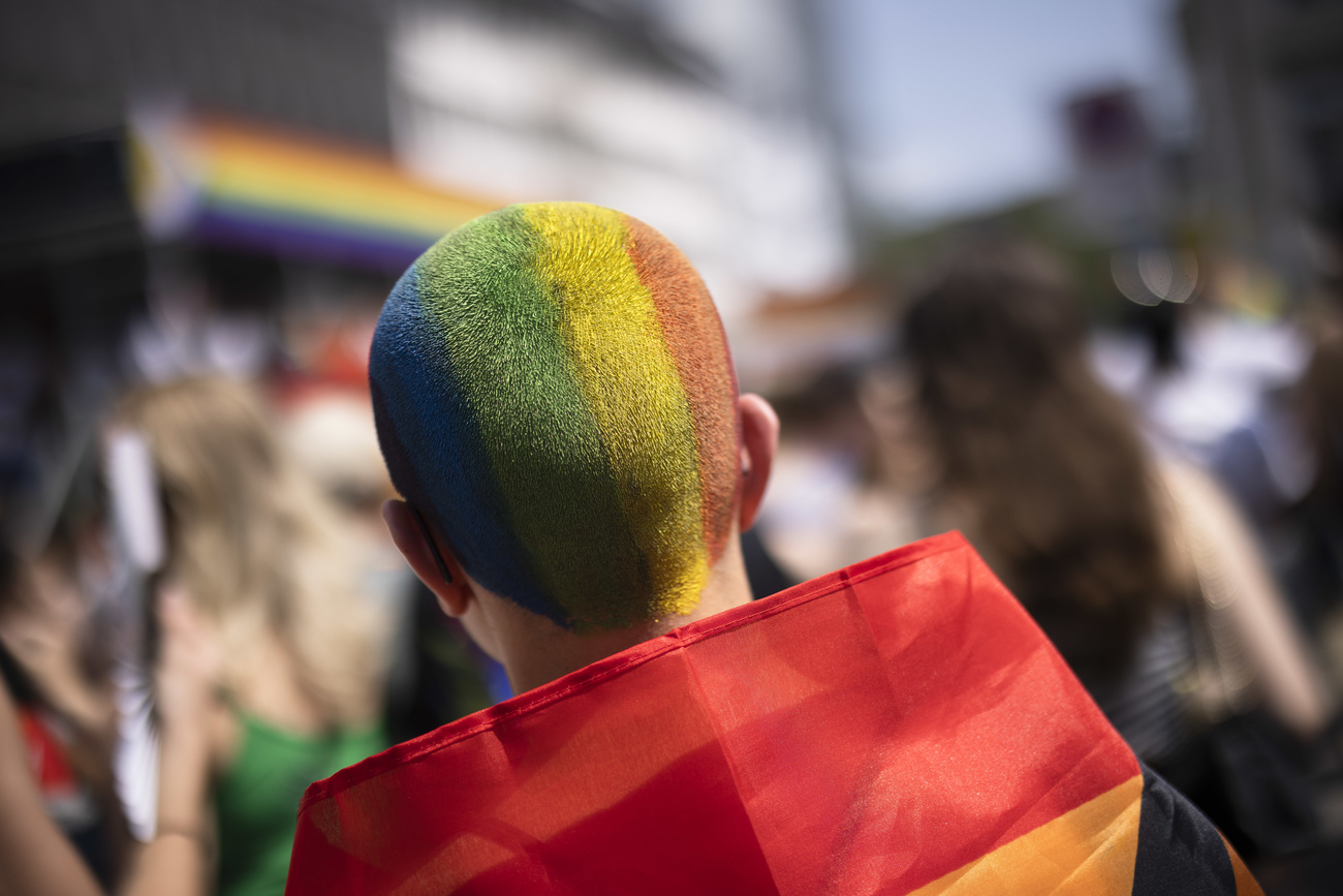 A person with a shaved head painted in rainbow stripes is shown with a rainbow flag draped around their shoulders.