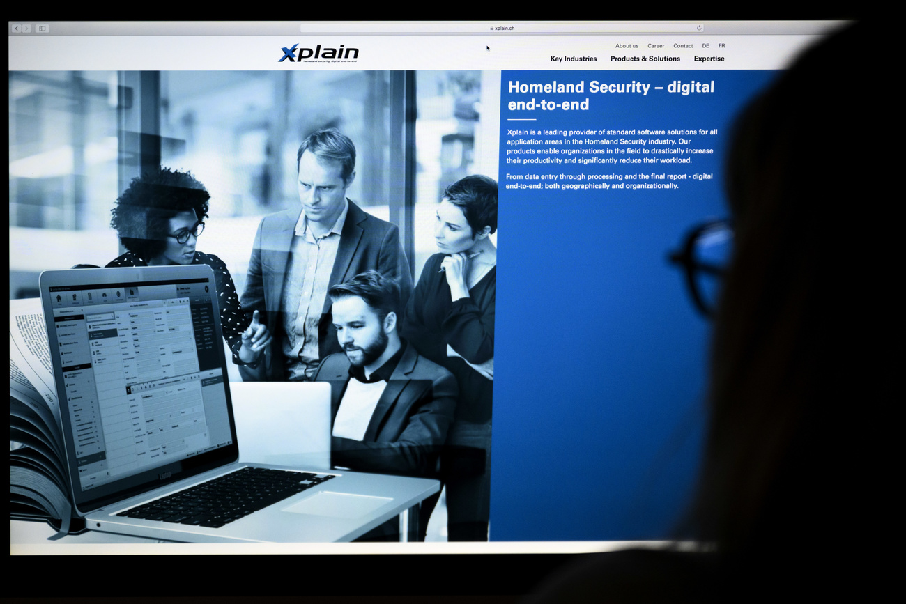 Photographed from behind, the dark silhouette of a person with glasses can be seen looking at a computer screen. On the screen is the Xplain website, which says ‘Homeland security – digital end-to-end' and has a picture of a group of four people gathered around a laptop.