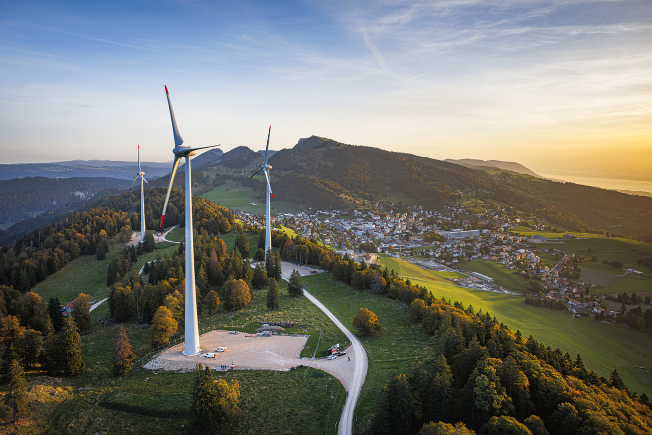 Three white wind turbines stand on a beautiful green mountainous landscape in Switzerland. They are surrounded by trees with a settlement visible in the background, along with mountain ranges and a blue sky to the left. The sun to the right is casting a golden glow on the mountains.