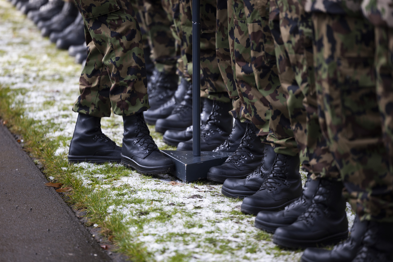 A line of legs from the knees down wearing the same army-issue camouflage trousers and black boots. The ground is grassy, with some tarmac visible to the left, covered with a light dusting of snow. One person’s feet can be standing facing the line of people and is using their foot to move a black metal stand.
