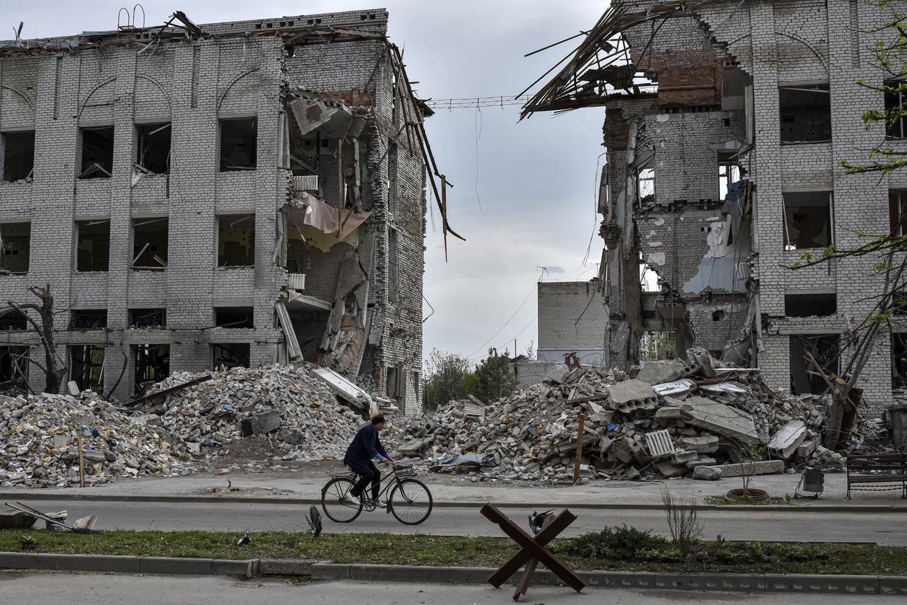 A woman rides a bicycle in front of a building destroyed by a Russian airstrike in the town of Orikhiv. Debris is piled up around the grey building.