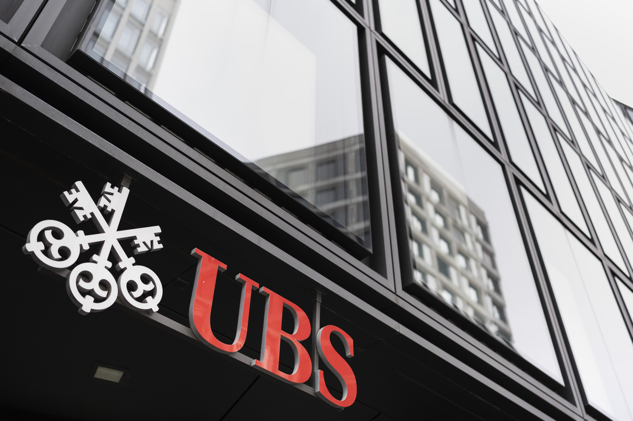 A picture of the red UBS lettering and interlocking three keys logo on a building. Other buildings are reflected in the building’s windows.