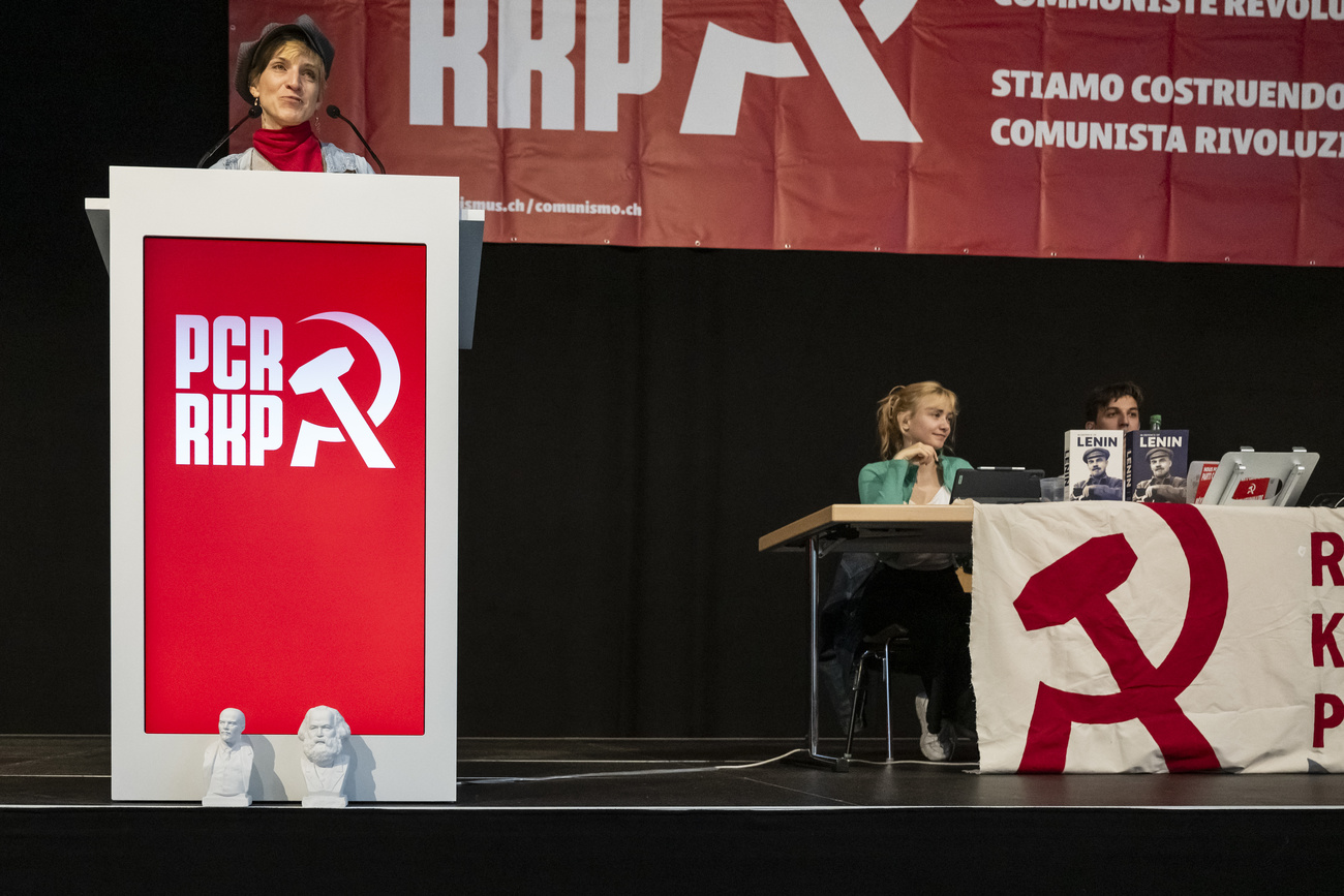 Olivia Eschmann of the Bern party section, left, speaks from behind a podium on a stage at the founding congress of the Revolutionary Communist Party. The red party logo is shown with its initials and the hammer and sickle symbol