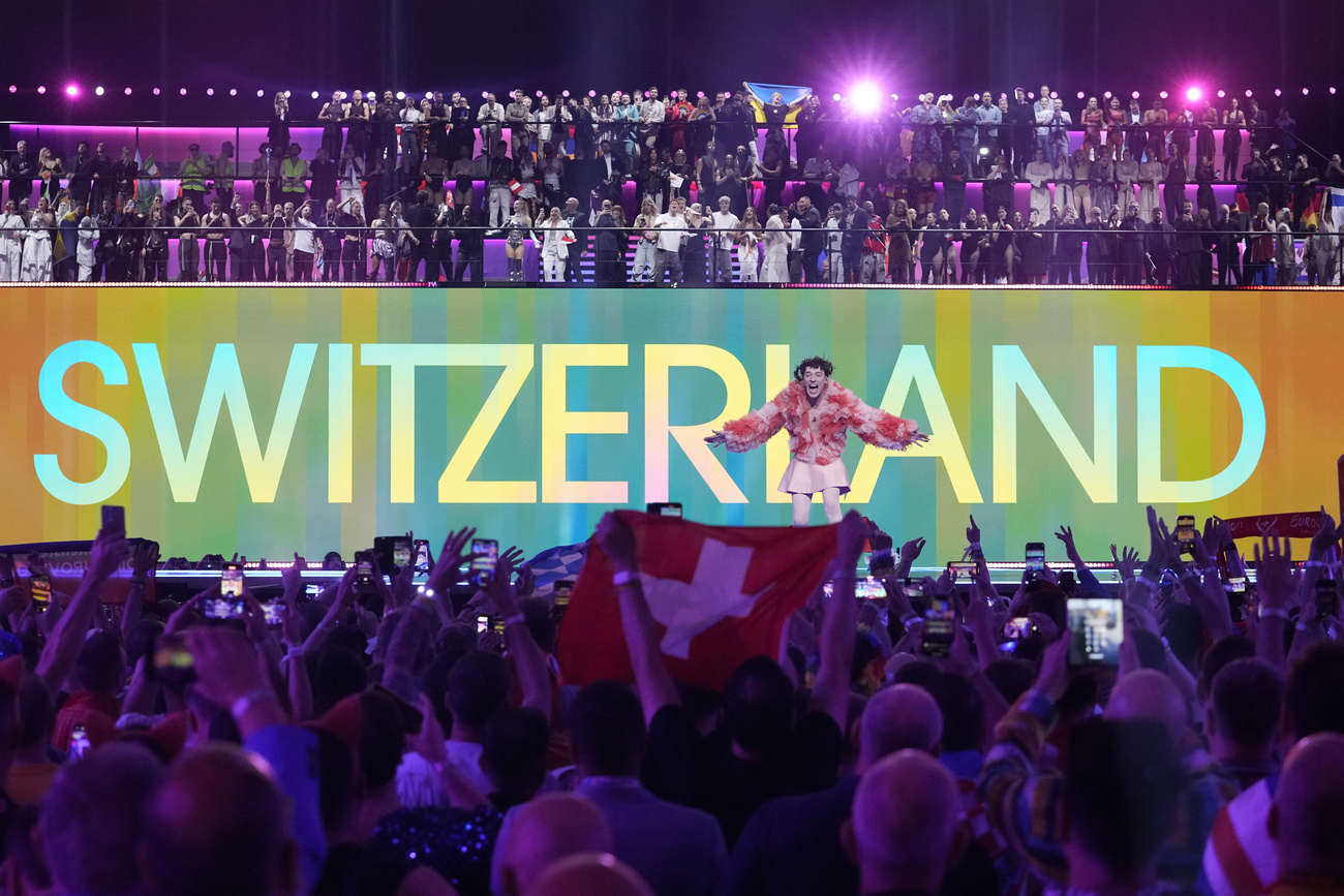 The singer Nemo in shown onstage at Eurovision 2024, while "Switzerland" is displayed behind.