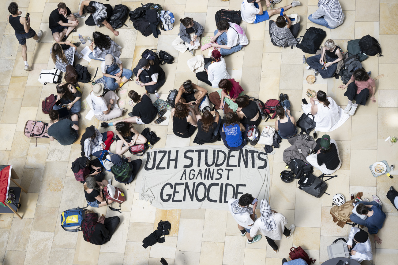 A group of students seated on the floor inside a university hall. In the middle of the group is a sign reading "UZH students against genocide".