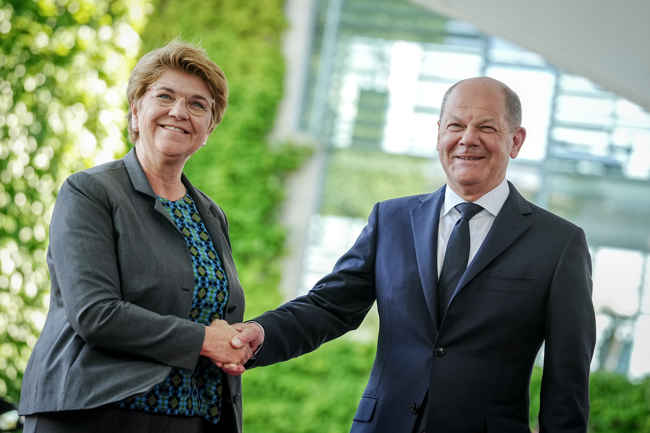 Swiss President Viola Amherd (left) and German Chancellor Olaf Scholz (right) shake hands and smile to the camera. Amherd is wearing a grey jacket and a blue and green printed top, Scholz wears a navy suit and tie with a white shirt.
