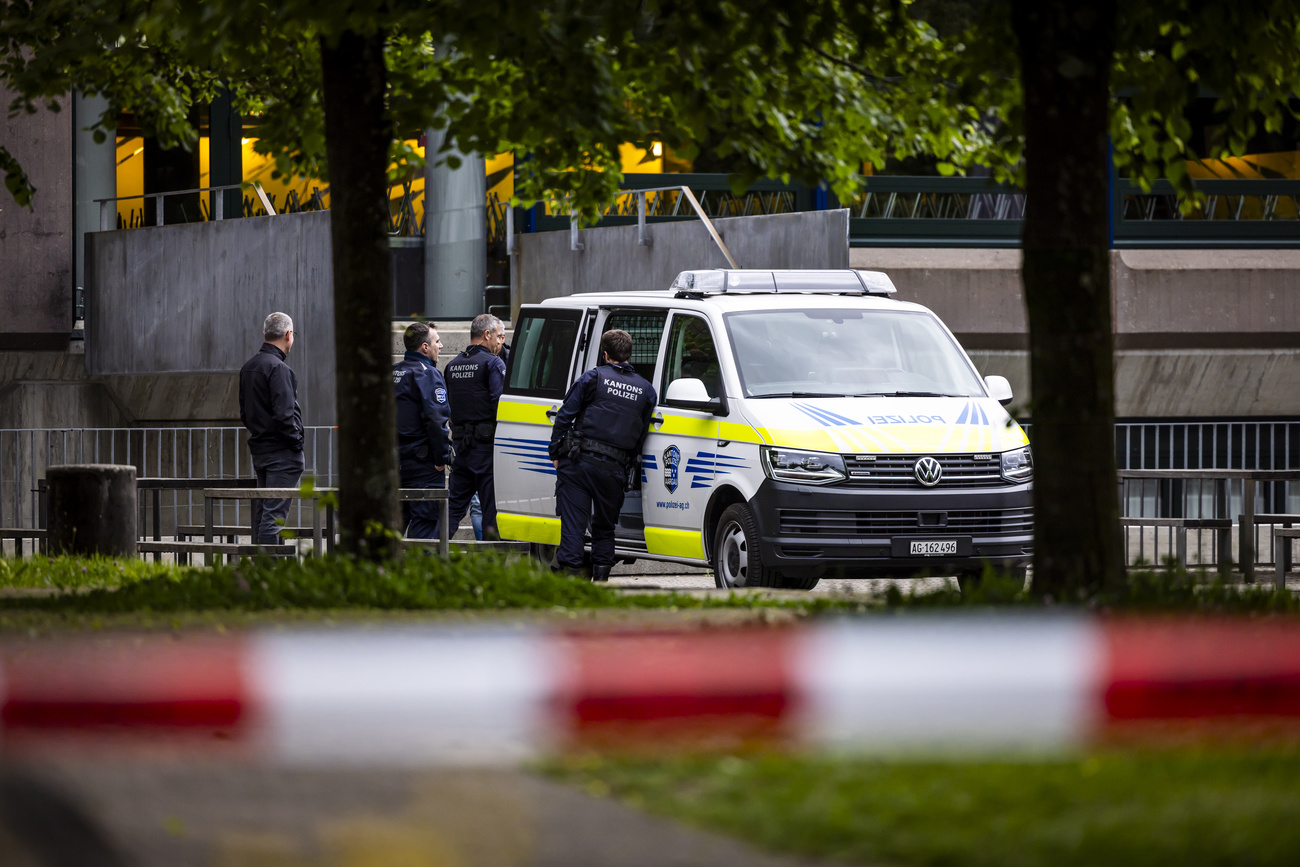 Across a red and white striped police line and a grassy area and in front of grey, concrete buildings can be seen a white police van with yellow and blue stripes. The door to the van is open and four male police officers in navy uniforms and black vests that say ‘cantonal police’ are standing next to the van.